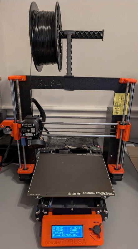 Prusa i3 MK3S+ years after assembly, sitting on project table. Spool of black filament on
   holder at top.